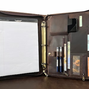 May include: Open brown leather portfolio with a zipper closure. The interior features a notepad, pen holders, card slots, and a pocket for a phone. The portfolio is open, revealing the contents, including a notepad with the text "TopFlight".