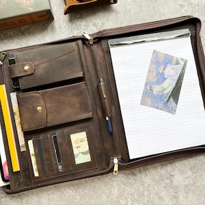 May include: A brown leather portfolio with multiple pockets and a zipper closure. The portfolio is open and has a notepad, a pen, and a card holder inside.