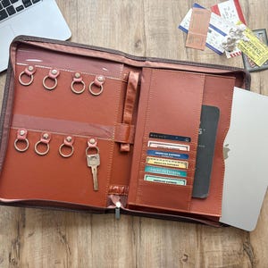 May include: A brown leather-like zippered portfolio organizer featuring eight metal key rings on the left side, one of which holds a silver key. The right side contains multiple card slots holding various credit cards and a vertical pocket for a notebook.