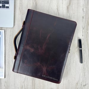 Puede incluir: Un portafolio de cuero marrón oscuro con asa, cierre y bolígrafo. El portafolio tiene una superficie texturizada y un logotipo en la parte inferior. Una computadora portátil y un periódico son parcialmente visibles en el fondo.