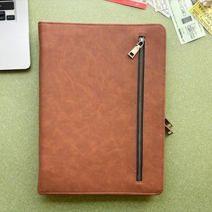 May include: A rectangular brown portfolio case with a textured surface and a vertical zipped pocket on the front. The zip features a metallic pull tab, and the case is constructed with visible stitching along the edges, providing a structured appearance for document storage.