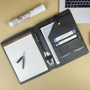 May include: An open, dark gray leather portfolio with a notepad, pens, and a white charging cable. The portfolio has pockets holding documents and tickets. A laptop and a white mouse are also on the light brown surface.