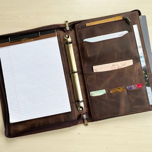 May include: A brown leather portfolio with a zipper closure, open to reveal a notepad and various pockets. The notepad has lined paper and the word "Veritas" printed on the top. The portfolio also has a three-ring binder.