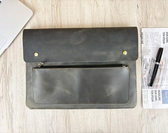 May include: A gray leather laptop sleeve with a front zippered pocket and a flap closure secured by two gold-colored studs. A pen and newspaper are next to the sleeve on a wooden surface. A laptop is partially visible.