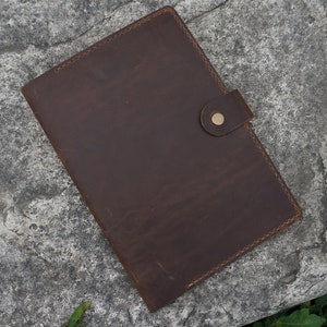 May include: A brown leather folder with a flap closure and a brass button. The folder has a stitched edge and is lying on a gray textured surface.