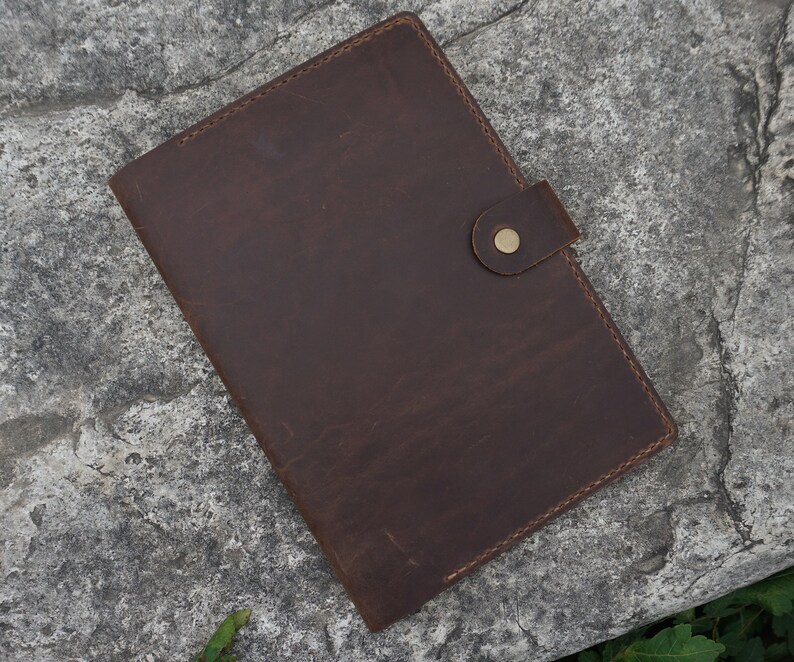 May include: A brown leather folder with a flap closure and a brass button. The folder has a stitched edge and is lying on a gray textured surface.