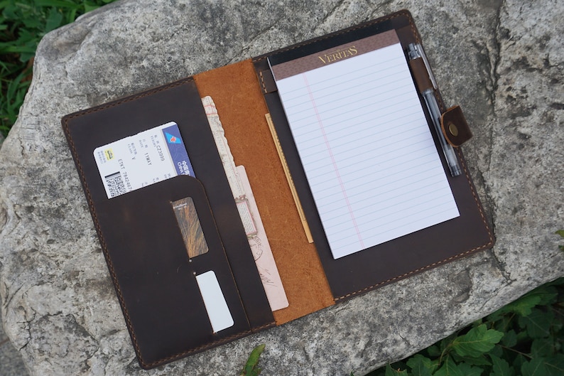 May include: A brown leather portfolio with a notepad, pen holder, and multiple pockets for holding documents and cards. The notepad has the word "Veritas" printed on the top.