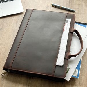 May include: A dark brown leather portfolio case with a zippered closure and a top handle. The front features a vertical slip pocket containing a newspaper with visible text. The portfolio is positioned next to a laptop and a pen.