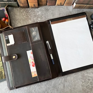 May include: A brown leather portfolio with a notepad, pen holder, and a flap closure. The portfolio has a lined notepad and a pen holder on the left side. The flap closure has a brass button.