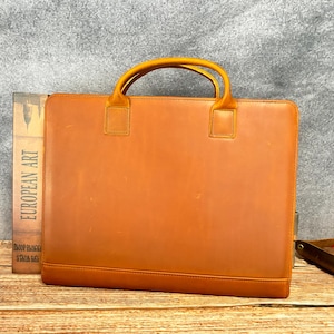 Può includere: Una ventiquattrore rettangolare in pelle marrone con maniglia superiore e base rettangolare. La ventiquattrore ha una superficie liscia e una maniglia marrone più chiara. Un libro con la scritta "EUROPEAN ART" è visibile a sinistra della ventiquattrore.