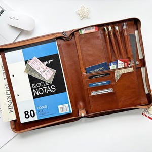 May include: A brown leather zippered portfolio organizer featuring an interior notepad labeled BLOC DE NOTAS, multiple card slots containing American Express cards and a passport, and four pen loops holding pens, all arranged in a structured layout.
