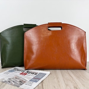 Puede incluir: Dos bolsos tote de cuero, uno marrón y otro verde oscuro, uno al lado del otro. Cada bolso tiene forma rectangular con la parte superior curvada y un asa. Un periódico está delante de los bolsos.