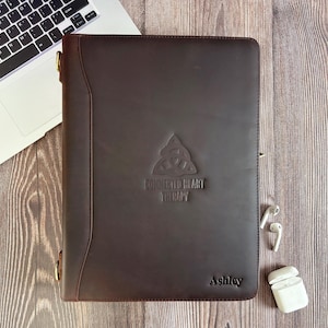 May include: A dark brown leather portfolio with the embossed text "CONNECTED HEART THERAPY" and the name "Ashley". The portfolio is next to a laptop and a pair of wireless earbuds.