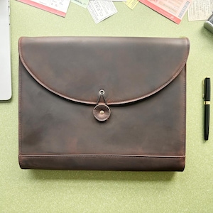 Puede incluir: Una carpeta para documentos de cuero marrón oscuro con un cierre de solapa grande y curvado, asegurado por un botón de cuero circular y un lazo de metal. La carpeta tiene un acabado mate suave y una forma rectangular con bordes cosidos abajo.