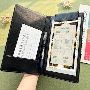 May include: Black leather menu holder with a menu, a pen, and a receipt. The menu is white with green and yellow accents and lists food and drink options. The receipt is white with black text. A blue pen is in the holder.