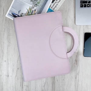 May include: A pale purple leather portfolio with a circular handle. A black smartphone and a laptop are visible. A magazine with text is also in the image. The portfolio is on a light wood grain surface.