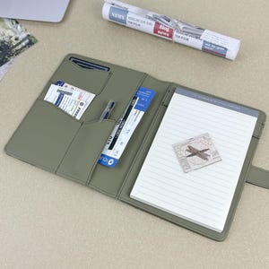 May include: An olive green leather travel organiser with a notepad, pen, and pockets holding tickets and a credit card. A rolled-up newspaper and a laptop are in the background. The notepad has a small airplane sticker.