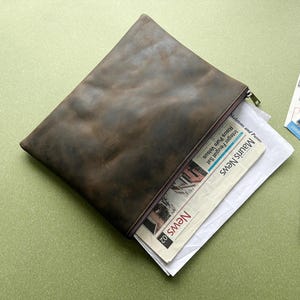 May include: A brown leather document case with a zip, partly open to show a newspaper. A passport, tickets, and two pens are placed beside it on a green surface. The newspaper is titled "Mauris News".
