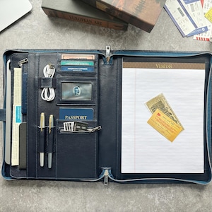 May include: A navy blue leather zippered portfolio organizer featuring multiple interior slots for credit cards, a passport, two pens, a coiled white charging cable, and a lined notepad with the word Veritas printed at the top.