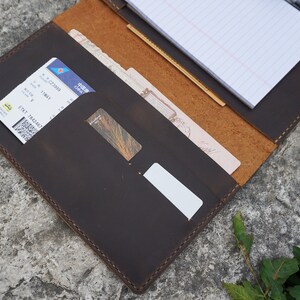 May include: A brown leather travel organizer with multiple pockets and slots for holding documents, cards, and a notepad. The organizer is open and shows a notepad with lined paper, a boarding pass, and a credit card.