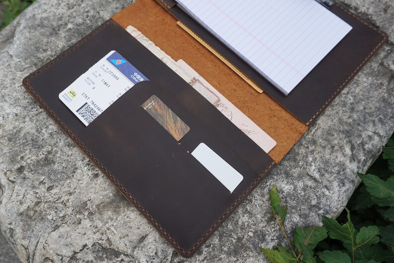 May include: A brown leather travel organizer with multiple pockets and slots for holding documents, cards, and a notepad. The organizer is open and shows a notepad with lined paper, a boarding pass, and a credit card.
