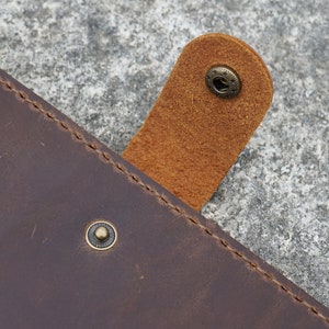 May include: Close-up of a brown leather journal with a flap closure. The flap has a brown leather strap with a brass button. The journal has a stitched edge and a brass button on the front cover.