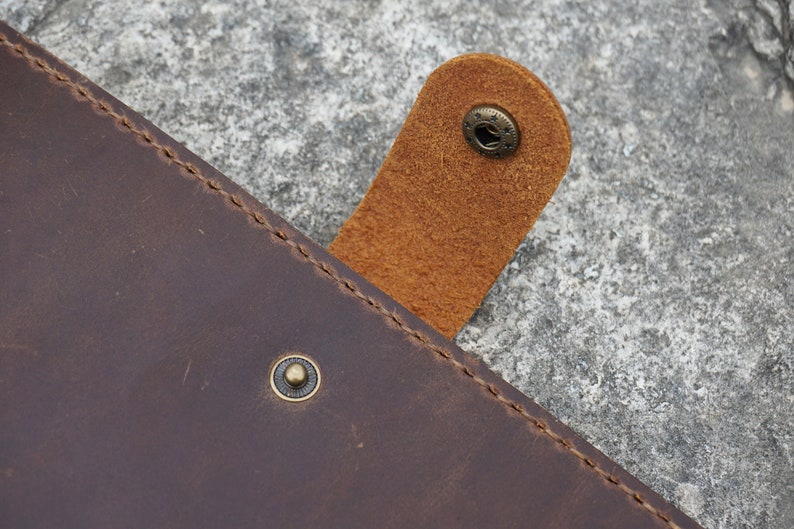 May include: Close-up of a brown leather journal with a flap closure. The flap has a brown leather strap with a brass button. The journal has a stitched edge and a brass button on the front cover.