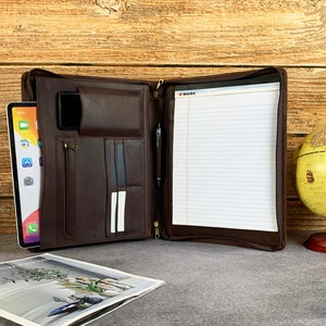 May include: A brown leather zippered portfolio with a notepad, pen, and a tablet inside. The portfolio has multiple pockets and slots for organization.