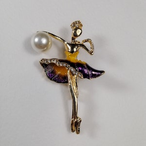 Puede incluir: Broche de bailarina dorada con falda morada y amarilla, un acento de perla y pedrería.