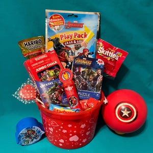 May include: A red bucket filled with Avengers-themed toys and treats. Includes a Play Pack, puzzle, stickers, bubble tube, chalk, candy, and a Captain America ball. The bucket is decorated with hearts.