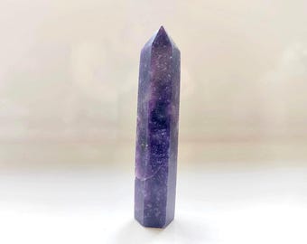 May include: A polished amethyst crystal tower with a hexagonal shape and a pointed top. The stone features deep purple hues with natural white inclusions and translucent areas throughout the vertical structure, standing upright on a flat base.
