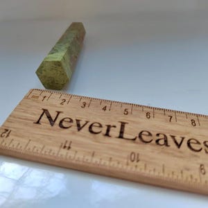 May include: A green and brown hexagonal crystal wand next to a wooden ruler. The ruler has the text "Never Leaves" engraved on it. The crystal wand is approximately 3 inches long.