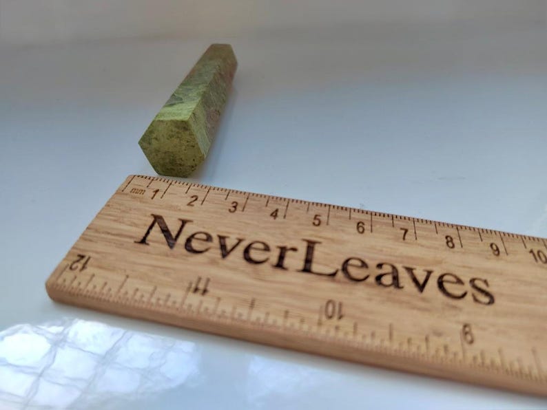 May include: A green and brown hexagonal crystal wand next to a wooden ruler. The ruler has the text "Never Leaves" engraved on it. The crystal wand is approximately 3 inches long.