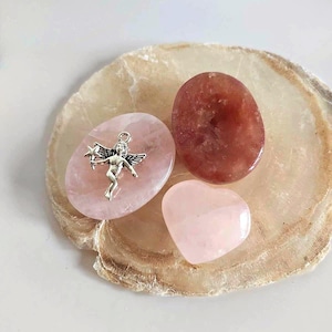 May include: A silver-toned metal angel charm with wings and a star wand rests on a smooth, oval-shaped light pink rose quartz stone. Beside it are a dark reddish-brown oval stone and a small, polished, heart-shaped light pink rose quartz stone.
