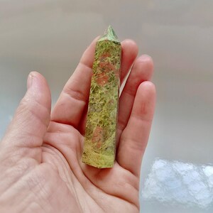 May include: A green and red unakite crystal tower. The polished stone has a rectangular shape with a pointed top. The unakite has a mottled pattern of green, pink, and orange hues. The crystal is held in a hand.