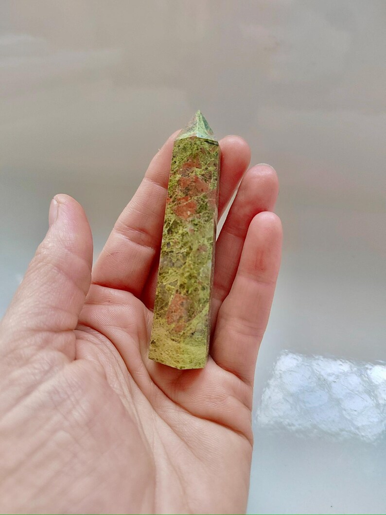 May include: A green and red unakite crystal tower. The polished stone has a rectangular shape with a pointed top. The unakite has a mottled pattern of green, pink, and orange hues. The crystal is held in a hand.