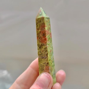May include: A polished unakite crystal obelisk. The stone features a mix of green, pink, and orange hues. The obelisk is held in a hand, showcasing its rectangular shape and pointed top. The background is a soft, blurred neutral color.