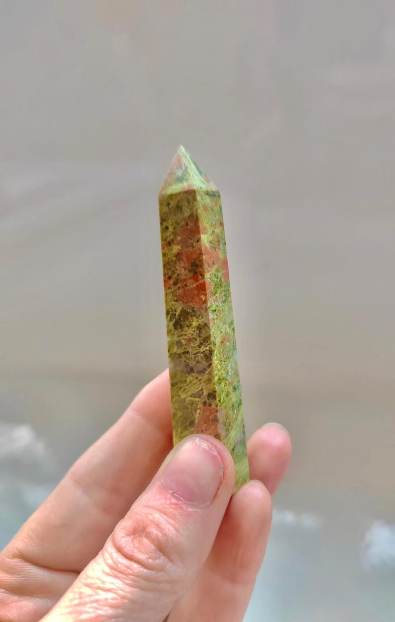 May include: A polished unakite crystal obelisk. The stone features a mix of green, pink, and orange hues. The obelisk is held in a hand, showcasing its rectangular shape and pointed top. The background is a soft, blurred neutral color.