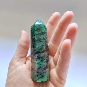 May include: A polished crystal tower featuring a hexagonal prism shape with a pointed top. The stone displays a mottled pattern of deep forest green, black, and vibrant magenta-pink inclusions throughout its smooth, multifaceted surface.