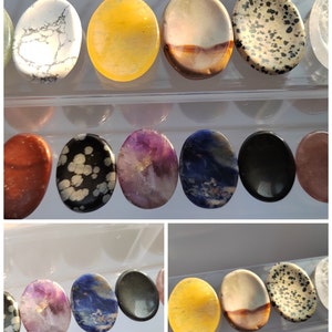 Pu&ograve; includere: Una collezione di pietre ovali levigate in vari colori e motivi, tra cui bianco, giallo, marrone, nero, viola, blu e rosso. Le pietre sono disposte in file su una superficie trasparente.