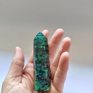 Puede incluir: Una torre de cristal de rubí zoisita pulida, sostenida en una mano. La piedra presenta una mezcla de tonos verdes, negros y rojos. La torre es rectangular con una parte superior puntiaguda, y los colores están arremolinados por toda la piedra.