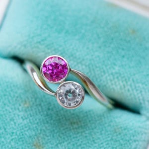 Puede incluir: Un anillo de plata con un diseño de derivación, con una gema redonda de color rosa brillante y una gema redonda transparente. El anillo se muestra en un joyero vintage de terciopelo azul claro.