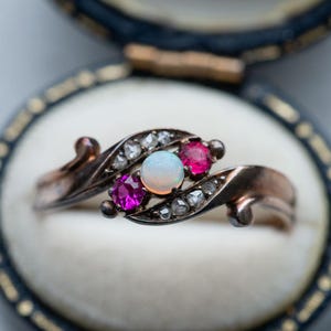 Puede incluir: Un anillo de estilo vintage con un diseño en espiral, engastado en metal oscuro. El anillo presenta una piedra de ópalo central, flanqueada por dos piedras rojas más pequeñas y acentuada con pequeñas piedras transparentes. El anillo se muestra en una caja de joyería negra y dorada.