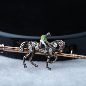 Puede incluir: Broche vintage con diseño de caballo y jinete. El caballo está adornado con piedras preciosas transparentes, y el jinete lleva un atuendo colorido. El broche tiene una barra dorada con gemas adicionales y un ojo de gema roja.
