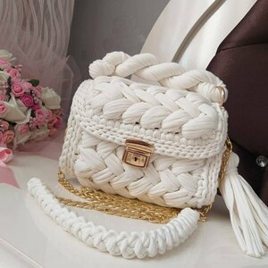 K&ouml;nnte beinhalten: Wei&szlig;e geh&auml;kelte Handtasche mit einer goldenen Kettenkette und einem Quastenanh&auml;nger. Die Tasche hat ein geflochtenes Design und einen goldenen Verschluss.