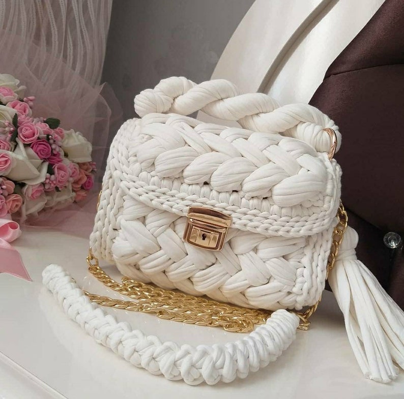 K&ouml;nnte beinhalten: Wei&szlig;e geh&auml;kelte Handtasche mit einer goldenen Kettenkette und einem Quastenanh&auml;nger. Die Tasche hat ein geflochtenes Design und einen goldenen Verschluss.