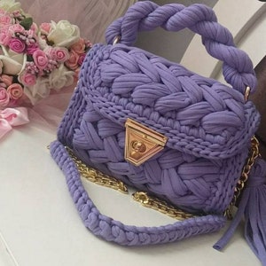 K&ouml;nnte beinhalten: Eine lavendelfarbene geh&auml;kelte Handtasche mit einer goldenen Kettenkette und einem Quastenanh&auml;nger. Die Tasche hat ein einzigartiges gewebtes Design und einen goldenen Verschluss.