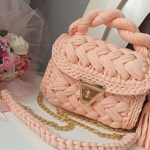 K&ouml;nnte beinhalten: Eine peachfarbene geh&auml;kelte Handtasche mit einer goldenen Kettenkette und einem Quastenanh&auml;nger. Die Tasche hat einen geflochtenen Henkel und einen goldenen Verschluss.