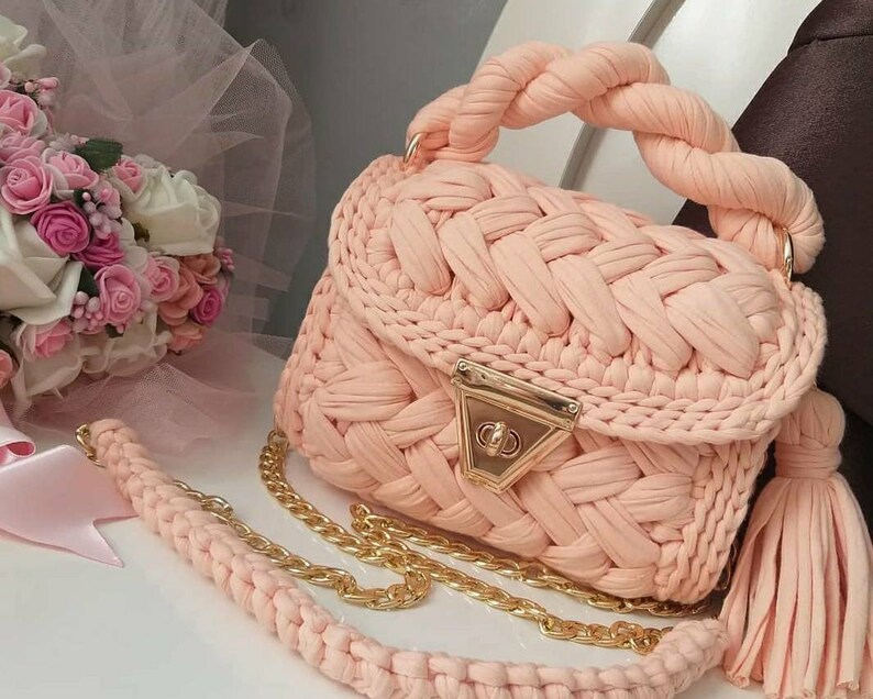 K&ouml;nnte beinhalten: Eine peachfarbene geh&auml;kelte Handtasche mit einer goldenen Kettenkette und einem Quastenanh&auml;nger. Die Tasche hat einen geflochtenen Henkel und einen goldenen Verschluss.