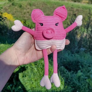 Könnte beinhalten: Ein rosa gehäkeltes Schweinchenspielzeug mit weißen Akzenten. Das Schweinchen hat lange Beine und eine herzförmige Nase.